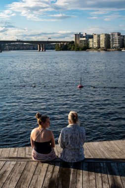Stockholm, İsveç 31 Temmuz 2024. İki kadın gün batımında, şehir merkezi ve Malaren Gölü manzaralı ahşap bir güvertede oturur..