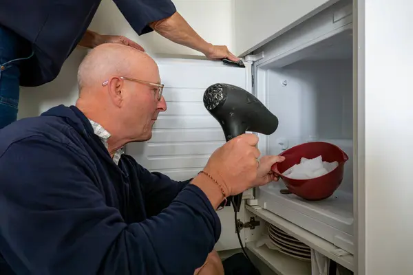 stock image A man defrosts a home kitchen freezer with a hair dryer to remove ice.