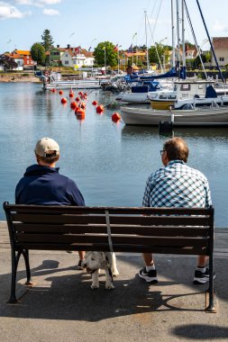 Oregrund, İsveç 18 Ağustos 2024 İki adam ve bir köpek limandaki bir bankta oturuyorlar.