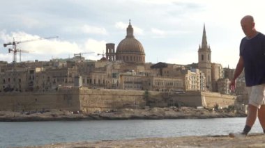 Valletta, Malta 'da bir erkek turist, Ball' s Bastion, St Elmo Bay ve Valletta 'nın Eski Şehir silueti üzerinde bir manzarayla kayalıklardan karşıya yürür..