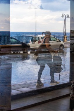 Sliema, Malta Nov 7, 2024 A woman walks on the sidewalk in the rain and seen through a window on the coastline. clipart