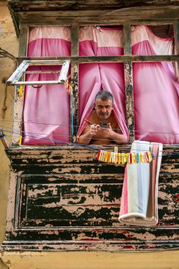 Valletta, Malta Nov 7, 2024 A man hangs out a window with pink curtains while smoking and checking his phone on St. Ursula Street. clipart