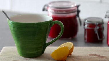 Kaynar su, ahşap bir tahtanın üzerine büyük bir fincana dökülüyor. Arka planda limon ve kavanoz dolusu taze çilek reçeli var.. 