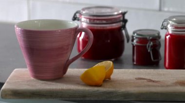 Kaynar su, ahşap bir tahtanın üzerine büyük bir fincana dökülüyor. Arka planda limon ve kavanoz dolusu taze çilek reçeli var.. 
