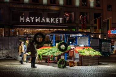 Stockholm, Sweden Dec 4, 2024 Pedestrians walk past a stand selling Christmas wreaths on Haymarket Square, or Hotorget, at night  . clipart