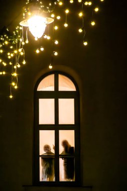 Riga, Latvia Dec 11, 2024 The silhouette of two people behind an arched window at night and Christmas decorations. clipart