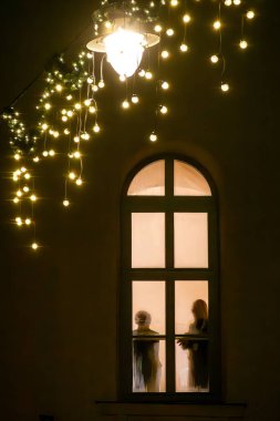 Riga, Latvia Dec 11, 2024 The silhouette of two people behind an arched window at night and Christmas decorations. clipart