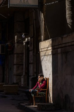 İskenderiye, Mısır 27 Ocak 2025 Bir adam bir iskan caddesinde bir güneş demetinde oturuyor telefonda.