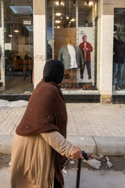 Alexandria, Egypt Jan 27, 2025 A senior woman with cane walks  on Al Naby Danyal, a fancy pedestrian street, past a modern men's clothing store. clipart