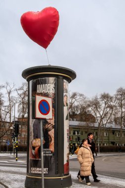 Stockholm, İsveç. 13 Şubat 2025. Yayalar Sevgililer Günü 'nde bir sokak büfesinin üzerinden uçan kırmızı bir kalp balonunun yanından geçiyorlar..