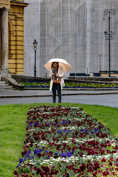 Zagreb, Hırvatistan 24 Mart 2025 'te Hırvat Ulusal Tiyatrosu' nun yanındaki parkta yağmurda şemsiyeli bir kadın ayakta duruyor.