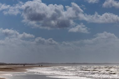 Skiveren, Danimarka Denizi, Skagen yakınlarındaki Skiveren sahilinde sörf ve bulutlar ve yürüyen insanlar..