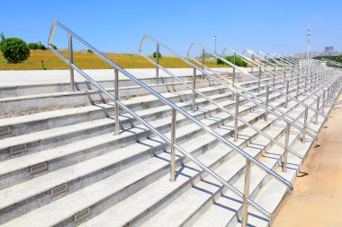 Açık mavi gökyüzünün altında beton merdivenler boyunca metal korkuluklar