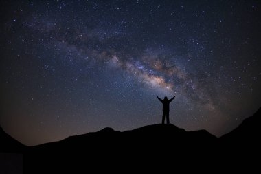 Samanyolu ile panorama manzarası, yıldızlı gece gökyüzü ve yüksek dağlarda kollarını kaldırmış ayakta duran sportif bir adamın silueti..