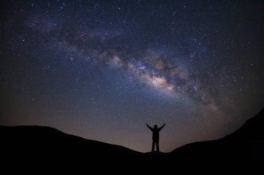 Samanyolu ile panorama manzarası, yıldızlı gece gökyüzü ve yüksek dağlarda kollarını kaldırmış ayakta duran sportif bir adamın silueti..
