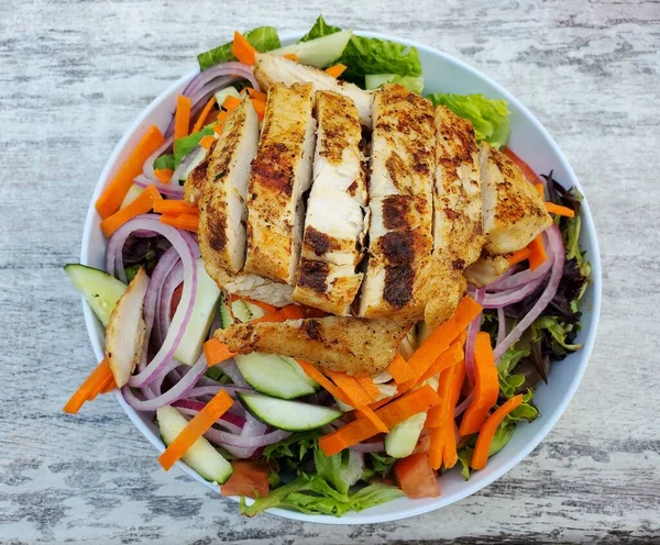 The top view of a garden salad with lettuce, cucumber, red onion, carrot, tomatoes, carrots with blackened chicken