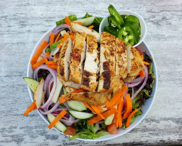 The top view of a garden salad with lettuce, cucumber, red onion, carrot, tomatoes, carrots with blackened chicken and a side of jalapeno slices