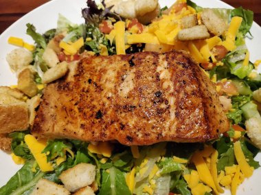 Salatanın üstünde ızgara somon, marul, kızarmış ekmek, peynir ve beyaz seramik tabakta sos.