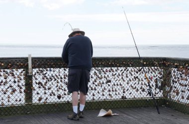 Okyanus Şehri, Maryland, ABD - 23 Mayıs 2025 - Bir balıkçı meraklısı Atlantik Okyanusu 'na bakan bir iskelede duruyor, sayısız asma kilitle süslenmiş bir tel örgüyle.