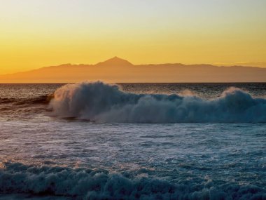 Agaete 'den (Gran Canaria) görülen Tenerife Kanarya Adası' nın siluetinin üzerinde altın gün batımı. Önünde daha yüksek bir püskürtme dalgası bulunan çalkantılı Atlantik..