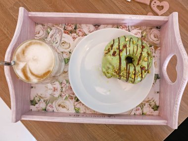 Bardakta kapuçinili bir tepsi ve yeşil fıstıklı bir donat ve çikolata şeritli beyaz bir tabak. Üst Manzara. Tablet masanın üstünde..