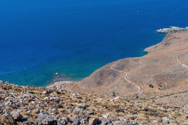 Sahildeki kayalık dağ yamacında yol ve mavi deniz suyuyla, güneşte, manzarada, Yunanistan, Girit adasında, telefoto