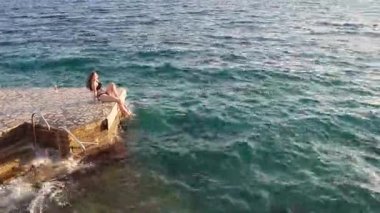 Siyah bikinili genç bir kadın denizin yumuşak dalgalarıyla çevrili kayalık bir sahil şeridinde oturuyor. Kamera onu yavaşça sağa dönmeden önce yakalıyor, engin denizi ve engebeli kıyı şeridini ortaya çıkarıyor, sakin ve sinematik bir atmosfer yaratıyor..