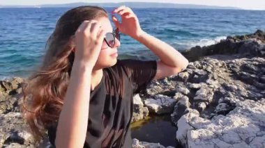 Güneş gözlüklü genç bir kadın kayalık bir kıyı şeridinde oturuyor ve uzaklığa bakıyor. Kamera, saçlarıyla oynayıp uçsuz bucaksız ufka bakarken yan profilini yakalıyor. Huzur, düşünce ve doğal güzellik anı..