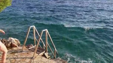 Metal bir merdivenden dikkatlice inen genç bir kadın kayalık bir kıyı şeridinden yumuşak bir şekilde dalgalanan denize giriyor. Etrafında deniz parıldıyor. Kendini ferahlatıcı suya daldırıyor. Doğanın güzelliğini kucaklıyor..