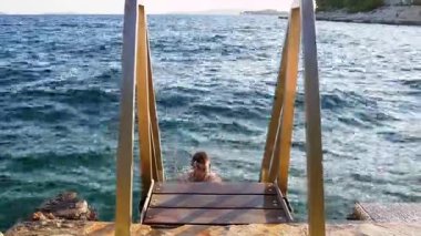 Genç bir adam, kayalık bir kıyı şeridine bağlı metal bir merdiveni tutarak nazikçe yuvarlanan okyanustan çıkar. Yükselirken etrafındaki dalgalar doğayla bir güç, macera ve bağ anını yakalıyor..