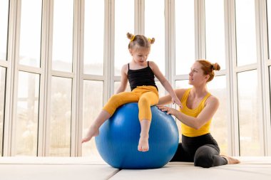 Young fit mom and her daughter having fun with fitness ball in a gym