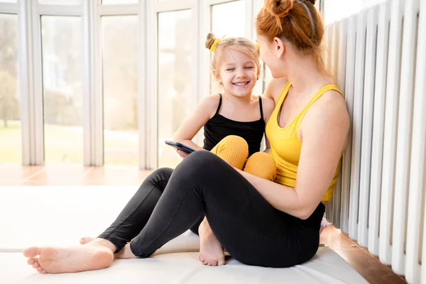 Young fit mom having loving time with her daughter, looking at a phone together