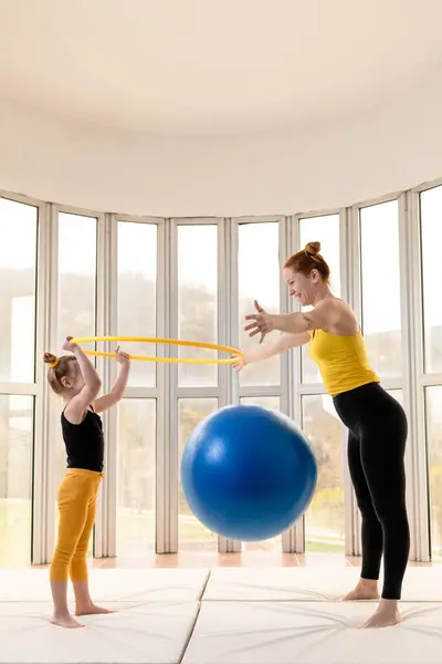 Young fit mom and her daughter having fun with hula hoop and fitness ball in a gym