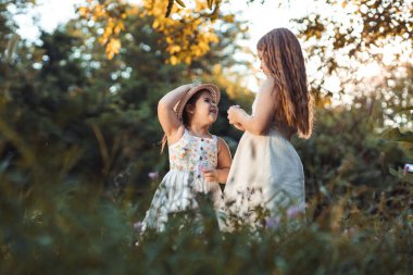 İki küçük kız, beyaz elbiseli kız kardeşler doğada çiçek topluyor.