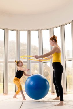 Young fit mom and her daughter having fun with hula hoop and fitness ball in a gym