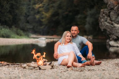 Nehir kenarında şenlik ateşinin yanında oturan genç çift. Boşluğu kopyala