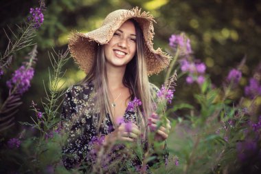 Mor çiçeklerle çevrili sarı şapkalı genç kız.