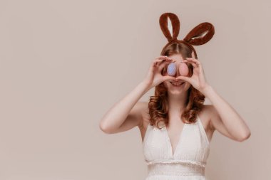 Beyaz elbiseli, tavşan kulaklı tavşan kulaklı, Paskalya yumurtalarını gözünün önünde tutan beyaz elbiseli, pastel bej arka planda duran beyaz bir kız.