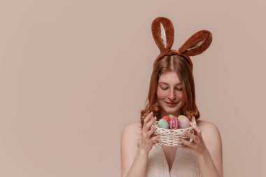 Tavşan kulaklı neşeli kız Paskalya sepetini tutuyor, aşağı bakıyor, yumuşak pastel arka plan, neşeli ruh hali