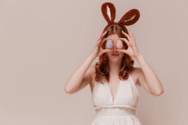 Tavşan kulaklı, beyaz elbiseli, neşeli bir şekilde gülümseyen, bahar kutlaması sırasında pastel bej arka planda duran tavşan kulaklı beyaz bir kız.