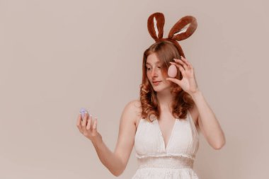 Tavşan Kulaklı Kızıl Kız elinde Paskalya yumurtası tutuyor ve bej arka planda onlara bakıyor.