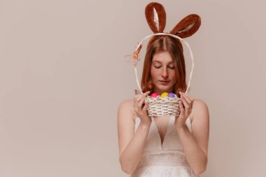 Tavşan kulaklı neşeli kız Paskalya sepetini tutuyor, aşağı bakıyor, yumuşak pastel arka plan, neşeli ruh hali