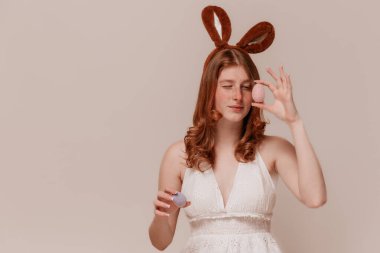 Tavşan Kulaklı Kızıl Kız elinde Paskalya yumurtası tutuyor ve bej arka planda onlara bakıyor.