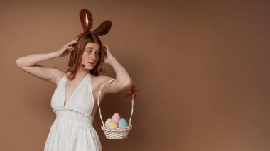 Beyaz elbiseli kızıl saçlı kız paskalya sepeti ve kahverengi arka planda tavşan kulaklı.