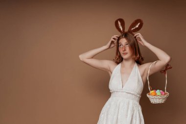 Beyaz elbiseli kızıl saçlı kız paskalya sepeti ve kahverengi arka planda tavşan kulaklı.