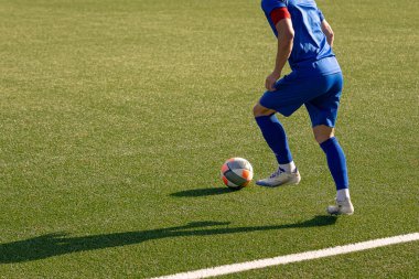 Mavi üniformalı futbolcu güneşli bir günde futbol sahasının kenarında top sürüyor..