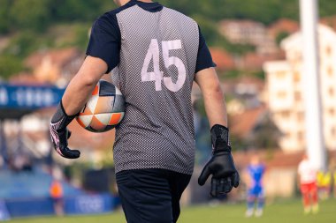 Sahada yürürken futbol topu tutan bir kalecinin arka görüntüsü, eldiven ve 45 numaralı forma giyiyor..