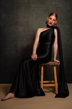 Siyah saten elbiseli zarif bir genç kadının tüm vücudu dikey bir portrede. Direk kameraya bakıyor, karamsar bir stüdyo ortamında poz veriyor..