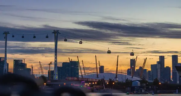 Gün batımında gökyüzünde süzülen teleferiklerle Londra ufuk çizgisinin nefes kesici görüntüsü. Bu manzaralı şehir manzarası, Greenwich, Londra 'da altın saatinde modern ulaşım ve mimari güzelliğin karışımını vurgular..