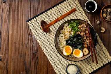 Geleneksel ramen ve domuz pastırması veya tavuk. Udon ya da ramen erişteli. Klasik kâselerde servis edilmiş. Arka planda Gyoza köftesi ve mantar var. Doğal ahşap zemin.